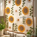 Crocheted kitchen curtain with sunflower and daisy patterns hangs in front of a window, casting light into the room, with a white sink and potted plants below.