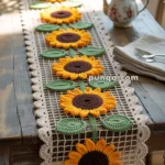 A table runner featuring a crocheted sunflower design with yellow petals and brown centers, accented by green leaves, placed on a wooden table alongside a decorative teapot and napkins with cutlery.
