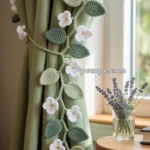 A green curtain is held back with a decorative crochet tie featuring green leaves and white flowers. A small bouquet of lavender in a glass vase sits on a nearby wooden table next to a book.
