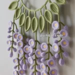 Crocheted wall hanging depicting lavender wisteria flowers with green leaves, suspended from a wooden rod.