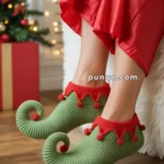 Person wearing elf-themed green slippers with curled toes and red details, sitting near a decorated Christmas tree and gift box.