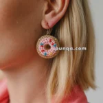 Close-up of a woman wearing a crochet earring shaped like a pink frosted donut with colorful sprinkles, paired with a pink outfit.