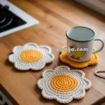 Crocheted flower-shaped coasters, two white with yellow centers and one plain yellow, on a wooden table with a mug.