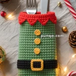 A crocheted cutlery holder resembling an elf's outfit, with green fabric, red scalloped trim, yellow buttons, and a black belt, holding a fork and knife, surrounded by festive Christmas decorations including pinecones, a candy cane, and string lights.