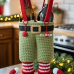Two wine bottles wrapped in a crochet holder shaped like elf pants, complete with suspenders, in a festive kitchen setting with holiday lights.