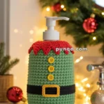 A soap dispenser covered with a crocheted Christmas elf costume, featuring red trim, yellow buttons, and a black belt with a yellow buckle, surrounded by festive holiday decorations including a garland and baubles.