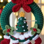 A crocheted Christmas wreath featuring a green circle with a red bow on top. Inside the wreath hangs a small crocheted Christmas tree decorated with white and red embellishments, surrounded by tiny stockings and leaves with red berries. It is displayed against a warmly lit background.