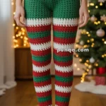 A person wearing festive crochet pants with a design resembling a Christmas elf, featuring a green belt section with a large buckle and red, green, and white striped legs, standing barefoot in a room with a decorated Christmas tree in the background.