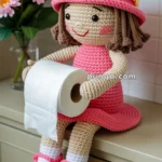 A crocheted doll with a pink dress and hat adorned with colorful flowers, holding a roll of toilet paper, sits on a bathroom cabinet next to a vase of pink flowers.