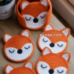 Crocheted orange and white fox-themed coasters with closed eyes, arranged around a matching crocheted basket with a fox design, on a wooden surface next to a plant.
