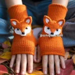 A pair of hands wearing orange knitted fingerless gloves with fox faces, surrounded by colorful autumn leaves on the ground.