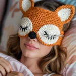 A person lying in bed wearing an orange crochet fox-themed sleep mask with closed eyes and ears.