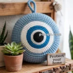 A crochet evil eye hanging decoration in blue and white colors is displayed on a wooden shelf next to a potted succulent and a small sign with the text "Protect your home."