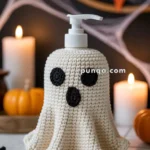 A soap dispenser covered with a knitted ghost-themed cover, featuring black crochet eyes and mouth, surrounded by Halloween decorations like small pumpkins, candles, and a spider, set on a tiled surface.