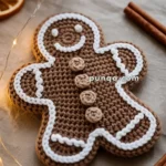 A crocheted gingerbread man with white icing details and three buttons is surrounded by festive decorations, including dried orange slices, star anise, and cinnamon sticks, accompanied by warm fairy lights.