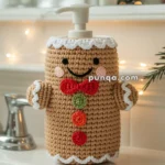 A soap dispenser covered with a crocheted cozy resembling a gingerbread man, complete with a red bow tie and colorful buttons, on a bathroom sink near a holiday-decorated background.