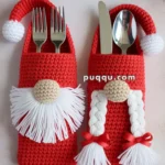 Two crocheted utensil holders designed like gnomes, with one featuring a white beard and the other with white braided hair, each holding forks and spoons. The holders are surrounded by Christmas decorations including pine cones, bells, and greenery.