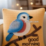 A knitted pillow featuring a colorful bird perched on a branch with the words 'good morning' stitched below, set against a yellow background.