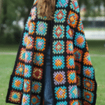 A person standing on grass wearing a colorful crochet patchwork blanket with a hood, featuring various geometric patterns in bright colors like blue, orange, and purple over a black base; they are also wearing blue jeans.