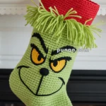 A Christmas stocking depicting a green, mischievous face with a furrowed brow, hanging from a fireplace mantel decorated with greenery and red ornaments. The stocking has a red cuff and is embroidered with notable facial features, resembling a fictional character.