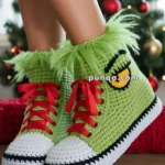 A pair of green crochet slippers with red laces and white soles, styled to resemble sneakers, being worn in front of a decorated Christmas tree with red and green ornaments.