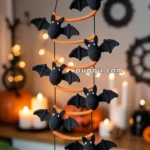 A handmade hanging decoration featuring a crescent moon and several crocheted black bats with orange accents, set against a blurred background with lit candles and pumpkins, creating a cozy Halloween atmosphere.