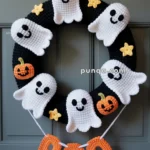 A Halloween wreath with crocheted white ghosts and orange pumpkins attached to a black circular base, with yellow stars scattered around. Hanging from the wreath are orange crocheted letters spelling "BOO."