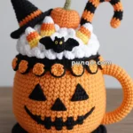 A crocheted Halloween-themed mug with a jack-o'-lantern face, topped with a witch hat, candy corn, a small pumpkin, and a bat. The mug also features black and orange striped details and a handle.
