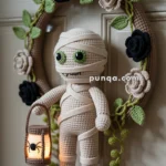 A crocheted doll of a mummy with large green eyes and a smiling face holding a small lantern adorned with a spider image. The doll is attached to a decorative wreath featuring crocheted black and beige roses with green leaves, hanging on a door.