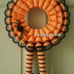 A Halloween-themed crocheted wreath featuring orange and black yarn with a lace edge, adorned with a crocheted spider. Hanging from it are crochet witch legs with orange and black striped stockings and black curled shoes with purple bows.
