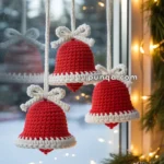 Three crocheted red bells with white trim hang in front of a window, adorned with white bows and surrounded by festive pine garland and lights.