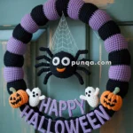 A crocheted Halloween wreath featuring alternating purple and black sections. A friendly crochet spider hangs from a web in the center, surrounded by small crochet pumpkins and ghosts. The words "Happy Halloween" are spelled out in purple letters at the bottom of the wreath.