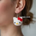 Close-up of a person wearing a crocheted earring shaped like a white cat with a red bow, with a website watermark reading "puqqu.com."