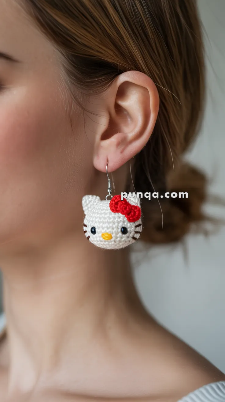 Close-up of a person wearing a crocheted earring shaped like a white cat with a red bow, with a website watermark reading "puqqu.com."
