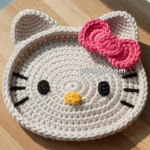A crocheted coaster shaped like a cat's face with black eyes, a yellow nose, pink bow, and whiskers, set on a wooden surface.