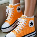 A pair of orange crochet sneakers with white laces and black star patterns, worn by a person sitting on a beige couch.