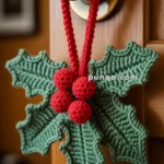 A decorative door hanger with green crocheted holly leaves and red berries hanging from a red cord on a brass doorknob.