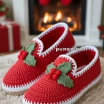 A pair of red crocheted slippers with green holly and red berries decoration, placed on a fuzzy rug in front of a fireplace with Christmas stockings and decorations.