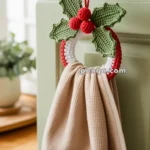 A beige kitchen towel hangs from a crocheted Christmas-themed ring adorned with green leaves and red berries, attached to a cabinet door handle.