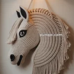A macrame wall hanging in the shape of a horse's head, with detailed woven features and fringe-like mane, illuminated by an overhead light.