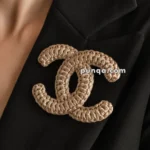 A gold crocheted brooch in the shape of intertwined letters, pinned to the lapel of a black jacket.