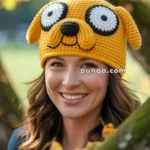 A woman smiling while wearing a crocheted hat resembling a cartoon character with large eyes and floppy ears, standing outdoors among autumn leaves.