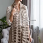 A woman standing indoors wearing a white blouse and beige pants, showcasing a crochet scarf with an open square pattern draped around her neck.