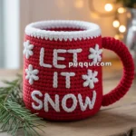 A red knitted mug with the words "LET IT SNOW" and white snowflake decorations, resting on a wooden surface with pine branches and blurred lights in the background.
