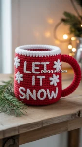 A red knitted mug with the words "LET IT SNOW" and white snowflake decorations, resting on a wooden surface with pine branches and blurred lights in the background.