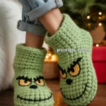 Person wearing green crochet slippers with a grinning face design, standing on a wooden floor near a Christmas tree with lights.