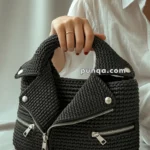 A person wearing a white shirt holds a stylish black woven handbag with zipper details and button accents, designed to resemble a biker jacket.