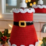 A soap dispenser on a bathroom countertop, decorated with a crocheted Santa outfit featuring a red body, white trim, and a black belt with a yellow buckle. In the background is a blurred Christmas tree with lights, and the countertop is adorned with holly leaves and candy canes.