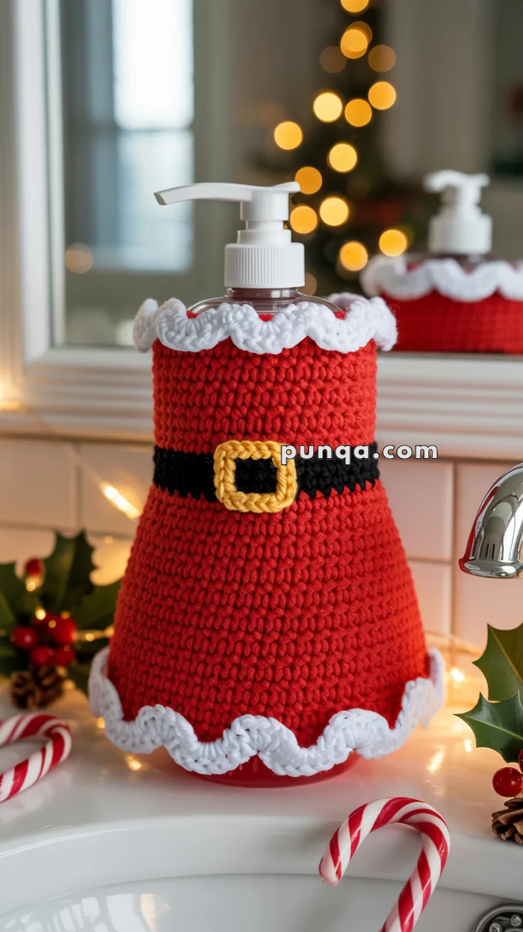 A soap dispenser on a bathroom countertop, decorated with a crocheted Santa outfit featuring a red body, white trim, and a black belt with a yellow buckle. In the background is a blurred Christmas tree with lights, and the countertop is adorned with holly leaves and candy canes.