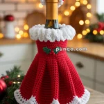 A wine bottle dressed in a red and white crocheted Santa outfit, featuring a skirt with a holly decoration, set against a festive kitchen background with Christmas lights.